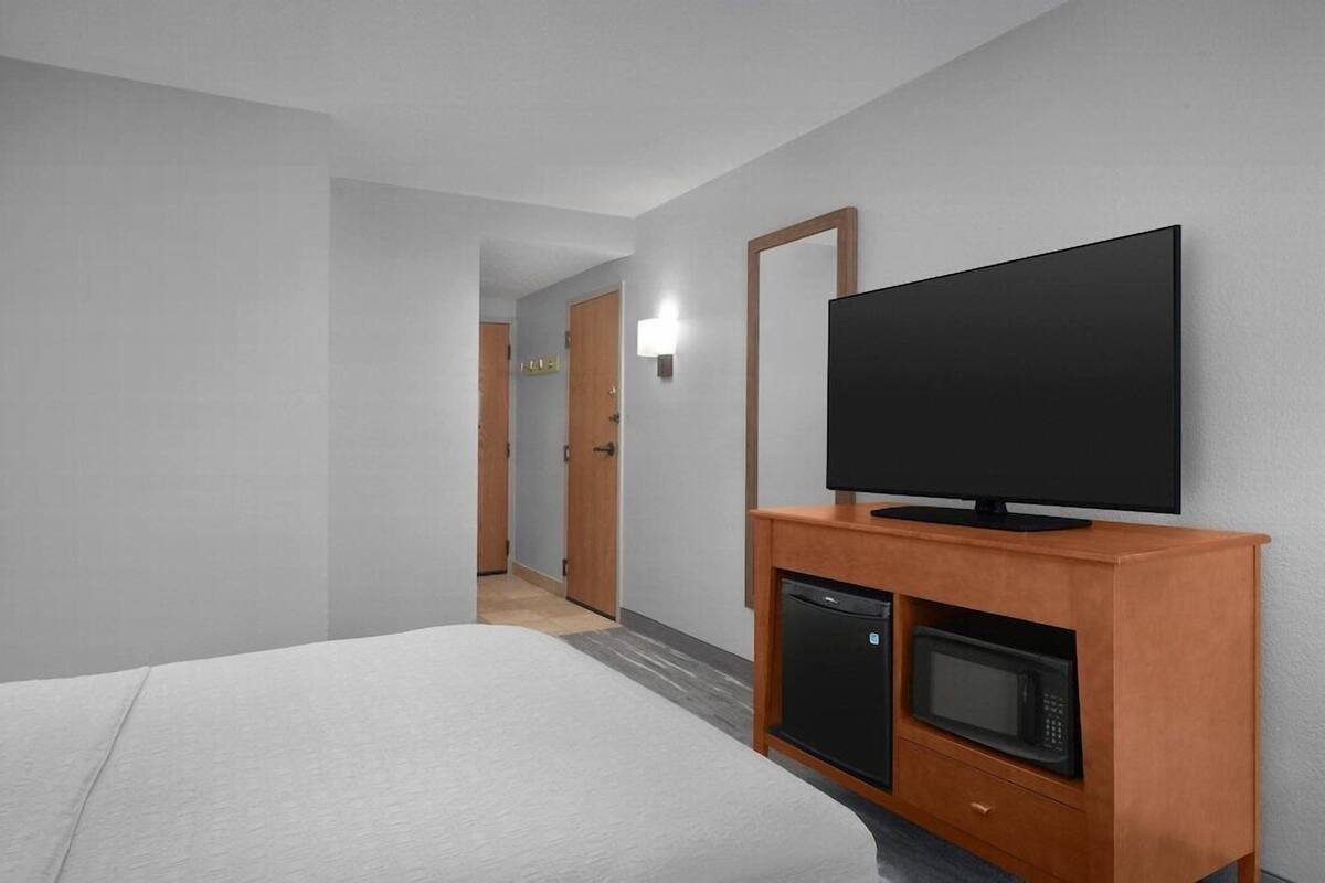 Une chambre d’hôtel avec un lit, un téléviseur posé sur une commode en bois, un four à micro-ondes et un couloir menant à une autre pièce.