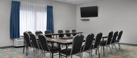 A conference room with a round table, chairs, a flat-screen TV, and a window with blue curtains.