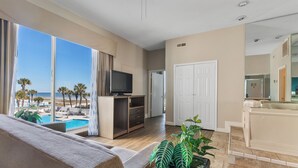 A modern living room with a large window overlooking a pool and beach, a flat-screen TV, a sofa, and a small table with plants. - Atlantic crest inn and suites (Myrtle Beach)