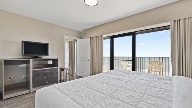 A hotel room with a large bed, a TV on a wooden desk, a balcony with a chair, and a view of the sea. - Atlantic crest inn and suites (Myrtle Beach)