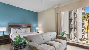 A hotel room with a large bed, a sofa, a nightstand with lamps, and a view of a building with balconies. - Atlantic crest inn and suites (Myrtle Beach)