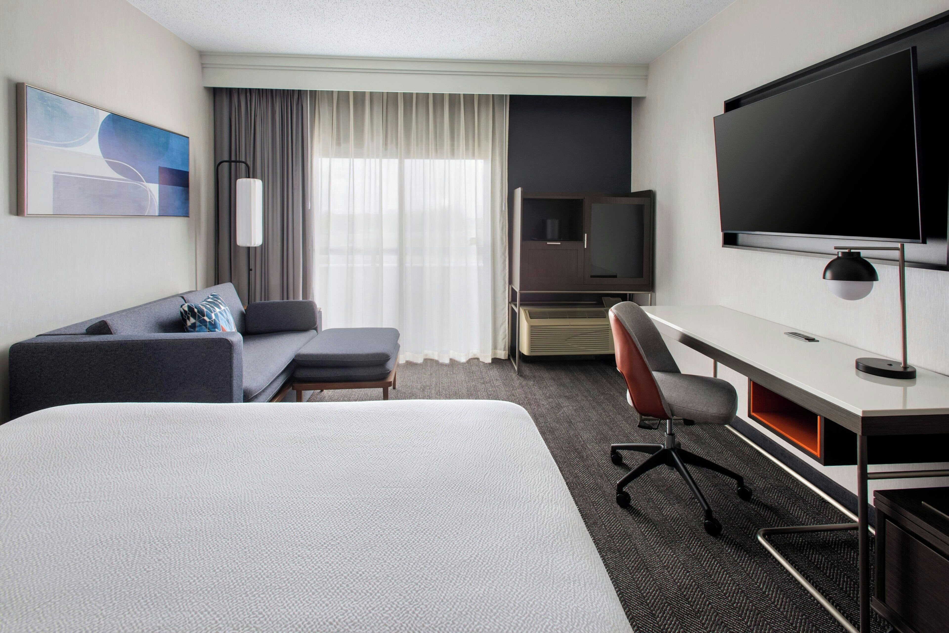 Habitación de hotel con cama, escritorio, silla, televisor, sofá y ventana con cortinas.