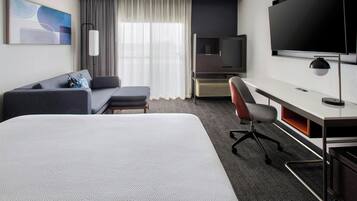Habitación de hotel con cama, escritorio, silla, televisor, sofá y ventana con cortinas.