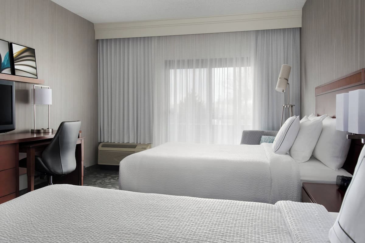 A hotel room with two beds, a desk, a chair, a TV, and a lamp.