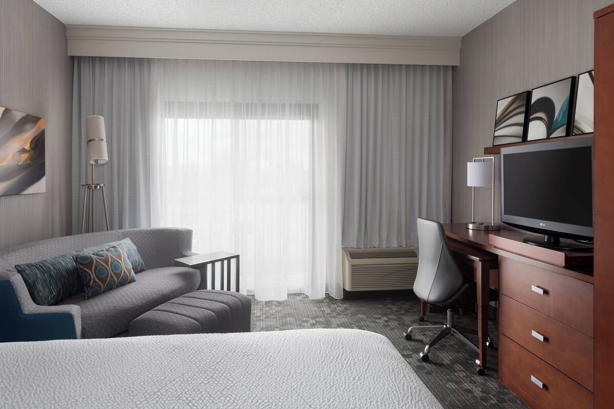 A hotel room with a bed, a desk with a TV, a chair, a couch, and a large window with curtains.