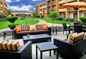 An outdoor seating area with a sofa, chairs, and tables, surrounded by a landscaped garden and a building in the background. - Courtyard Syracuse Carrier Circle (East Syracuse)