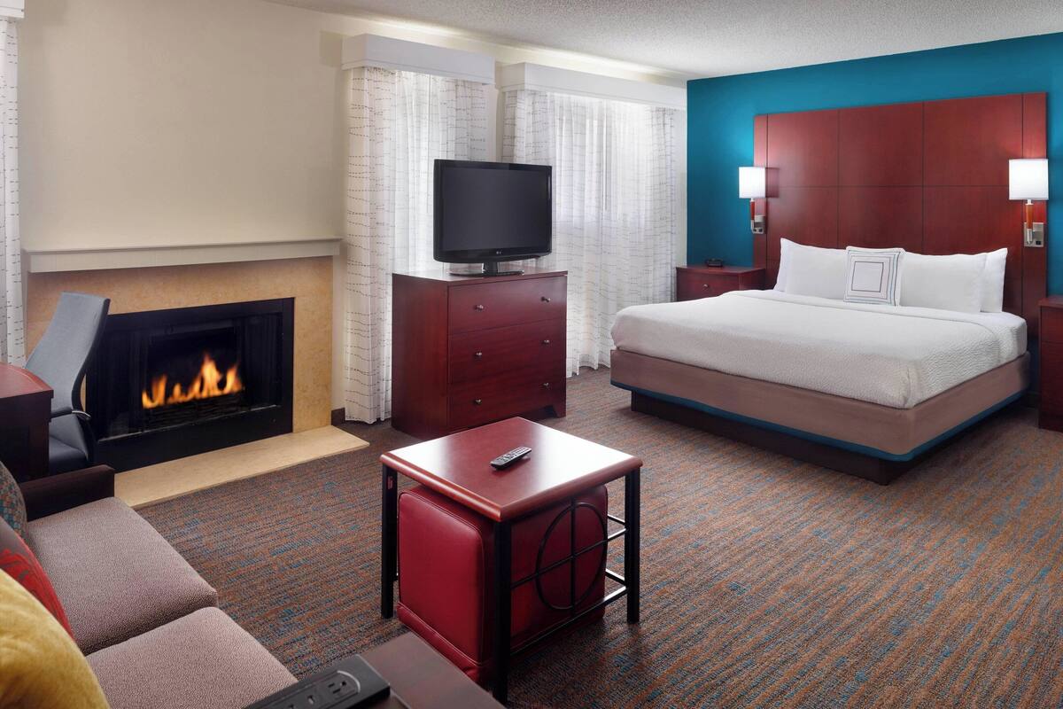 Une chambre d’hôtel avec une cheminée, un lit, un téléviseur posé sur une commode en bois, une table de chevet rouge et un mur d’accent bleu.