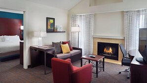 Una habitación de hotel con cama, sofá, sillón rojo, mesita, televisión, chimenea y lámpara.