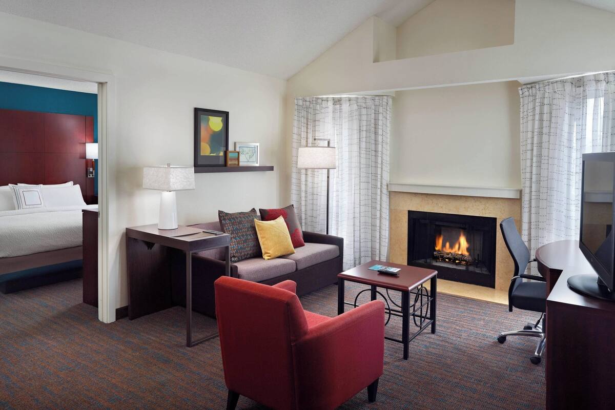 Une chambre d’hôtel avec un lit, un canapé, un fauteuil rouge, une petite table, une télévision, une cheminée et une lampe.