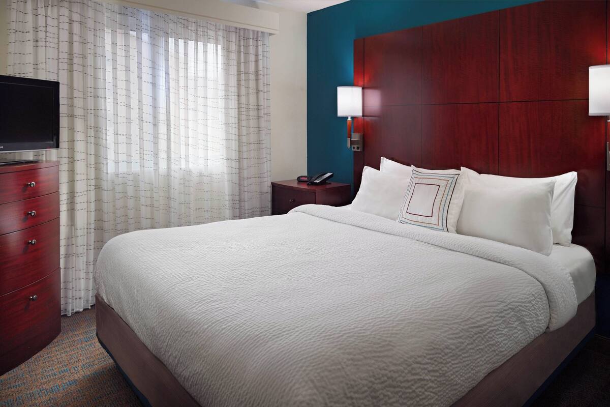 Une chambre d’hôtel avec un grand lit, un téléviseur posé sur une commode en bois, une table de chevet avec une lampe et une fenêtre avec des rideaux.
