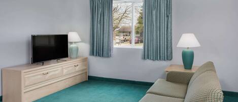 Una habitación con televisor sobre un mueble de madera, un sofá, una ventana con cortinas y una lámpara en una mesita de noche.