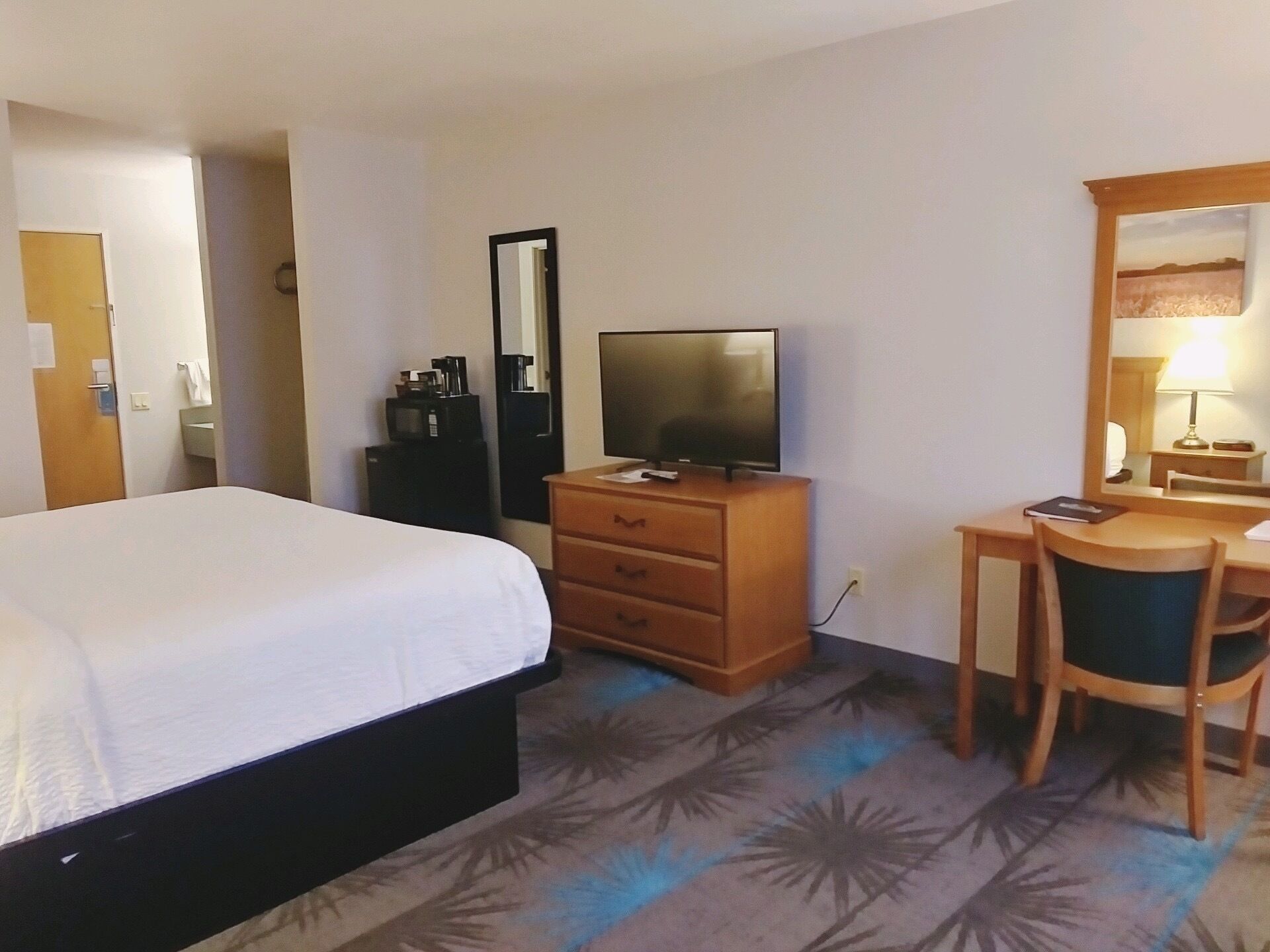 Une chambre d’hôtel avec un lit, un téléviseur posé sur une commode en bois, un bureau avec une chaise et un miroir.