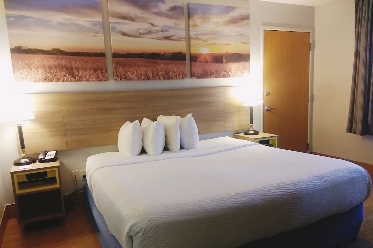 Ein Hotelzimmer mit einem großen Bett, zwei Nachttischen, einer Lampe, einem Gemälde, das einen Sonnenuntergang über einem Feld zeigt, und einer Holztür.