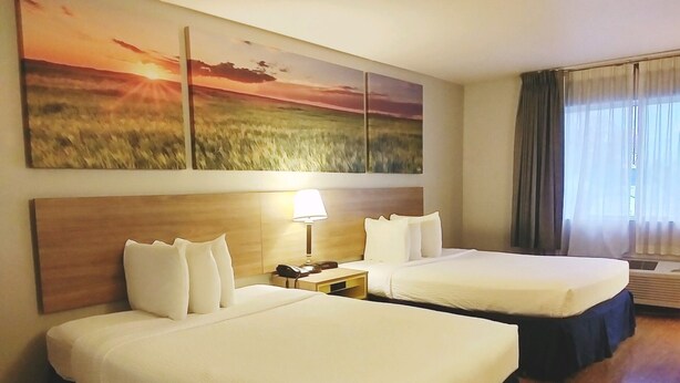 A hotel room with two beds, a nightstand with a lamp, a painting of a sunset, and a window with curtains.