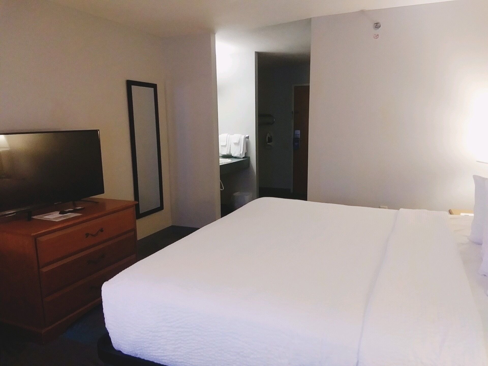A hotel room with a large bed, a wooden dresser with a flat-screen TV, a full-length mirror, and a door leading to a bathroom.