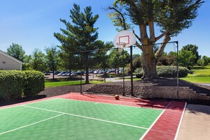 Una cancha de baloncesto al aire libre con aro, rodeada de árboles y un estacionamiento.