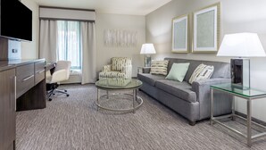 A hotel room with a grey sofa, a round glass coffee table, a grey armchair, a TV on a wooden cabinet, and two lamps on side tables.