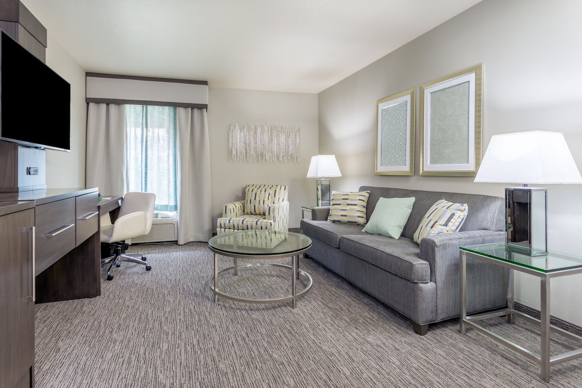 A hotel room with a grey sofa, a round glass coffee table, a grey armchair, a TV on a wooden cabinet, and two lamps on side tables.