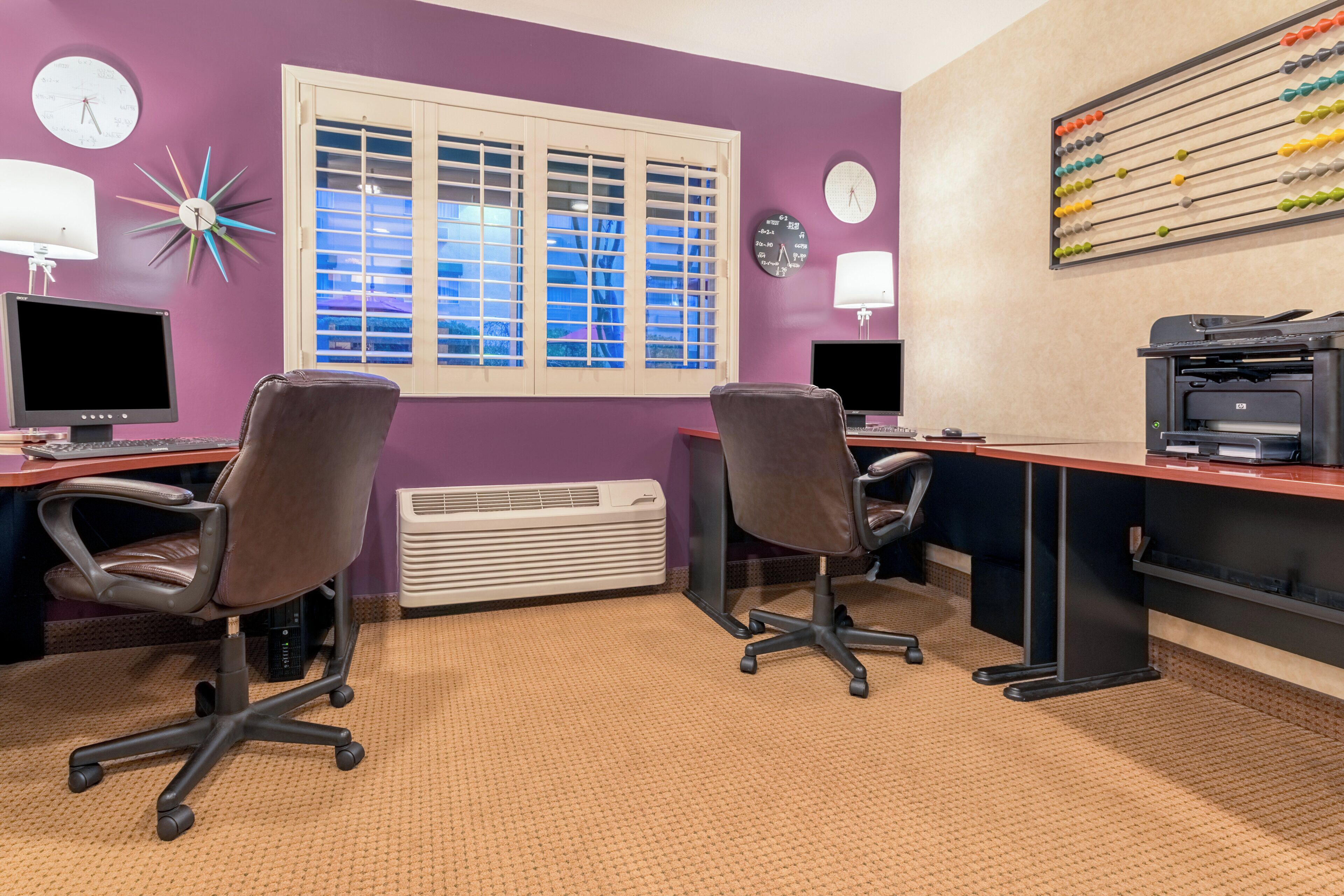 Quarto com duas mesas, cadeiras, computadores, uma impressora e janela com persianas.