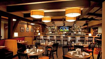 A restaurant with multiple tables and chairs, a bar area with a television, and a brick wall with framed pictures.
