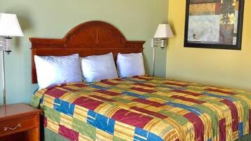 A hotel room with a wooden headboard, a colorful quilted bedspread, two bedside lamps, and a framed artwork on the wall.