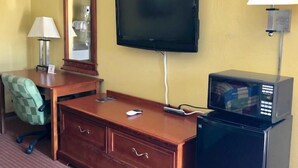 Habitación de hotel con escritorio de madera, televisor de pantalla plana, microondas, lámpara, silla y cómoda.
