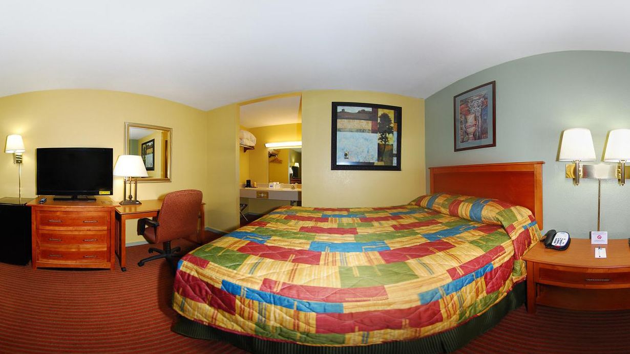 A hotel room with a large bed, a desk with a television, a chair, a desk lamp, a nightstand with a phone and a sign, and a painting on the wall.