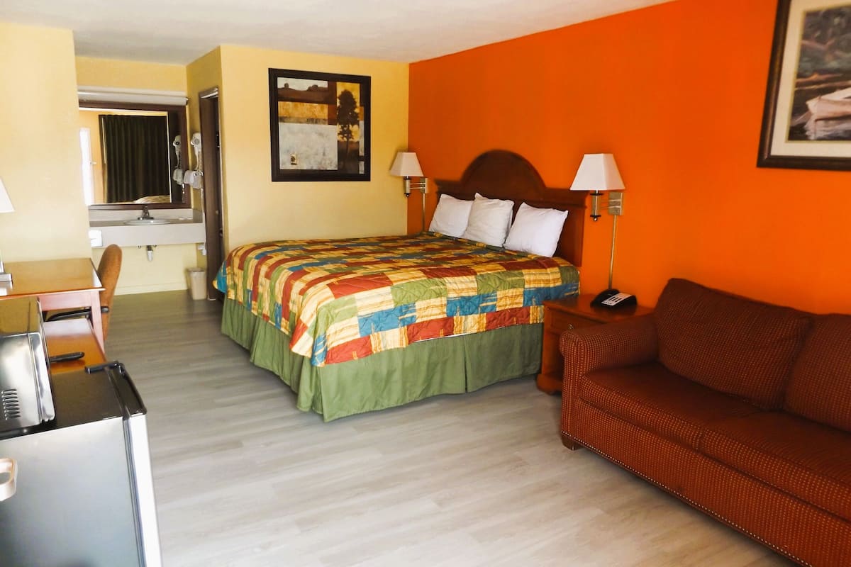 A hotel room with a bed, a sofa, a desk with a chair, a lamp, and a painting on the wall.