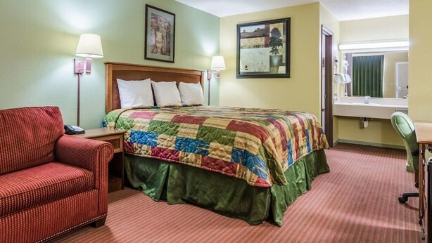 Habitación de hotel con una cama, un sillón rojo, un escritorio con una silla y una ventana con vista a un edificio.