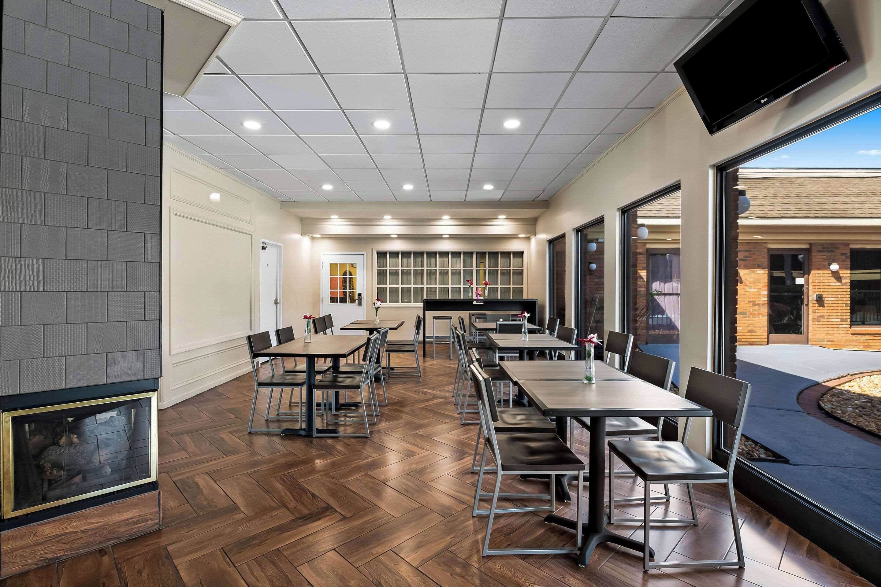 Un espace repas moderne avec un plancher en bois, des chaises et des tables en métal. Une cheminée se trouve à gauche et de grandes fenêtres à droite.