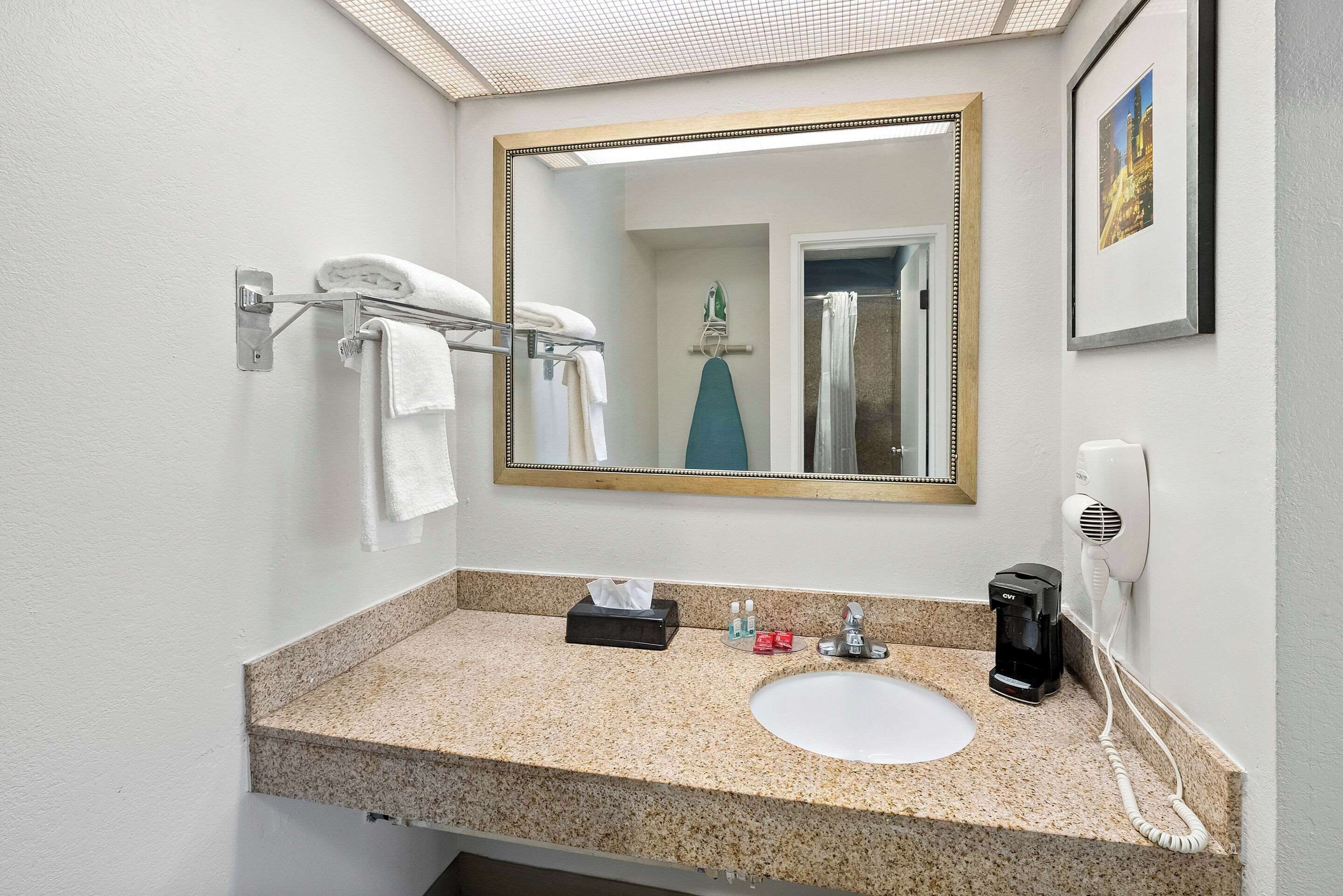 A bathroom with a granite countertop, a white sink, a mirror with a wooden frame, a towel rack with white towels, a tissue box, a hairdryer, and a framed picture on the wall.