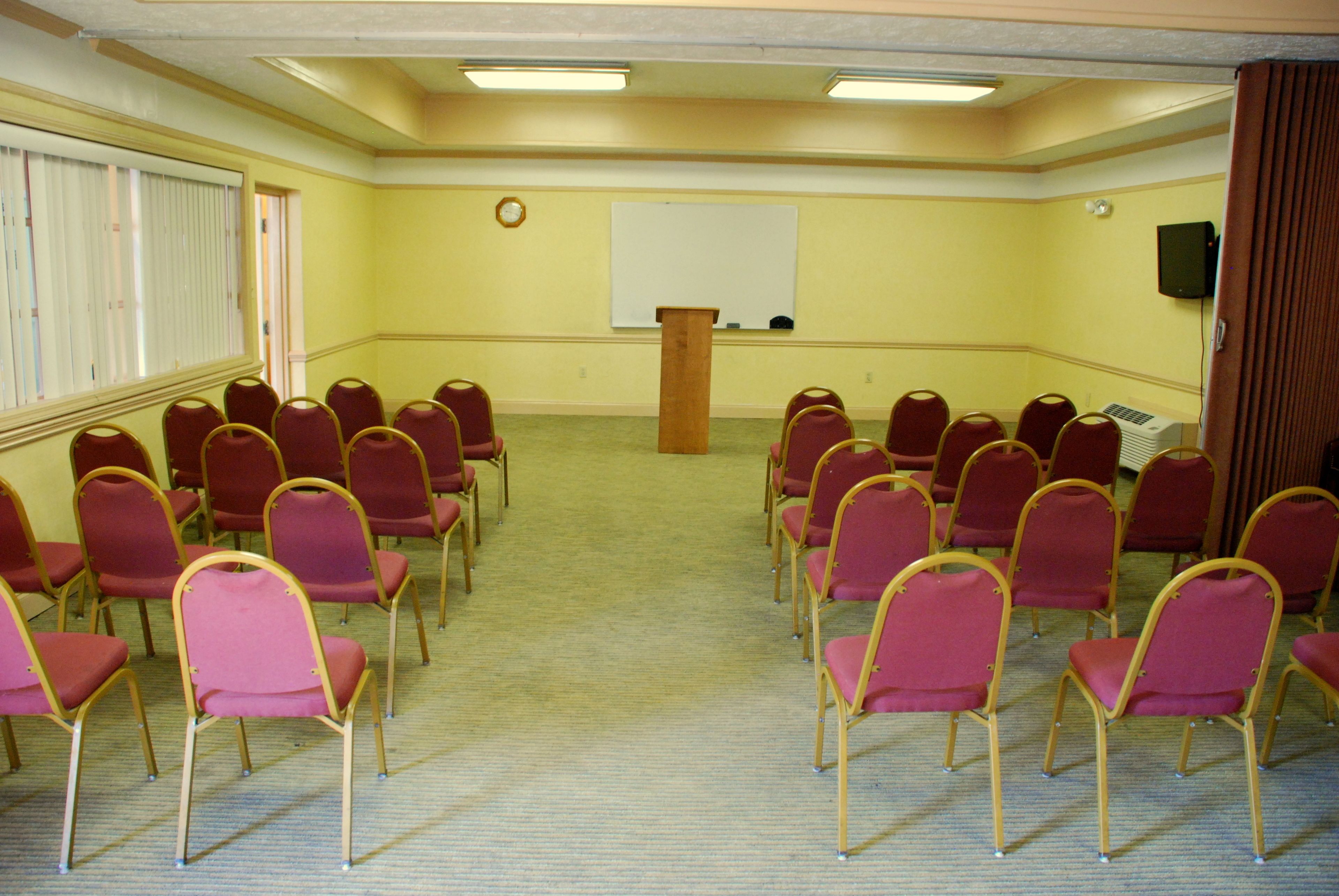 A conference room with rows of chairs, a podium, a whiteboard, and a clock on the wall.