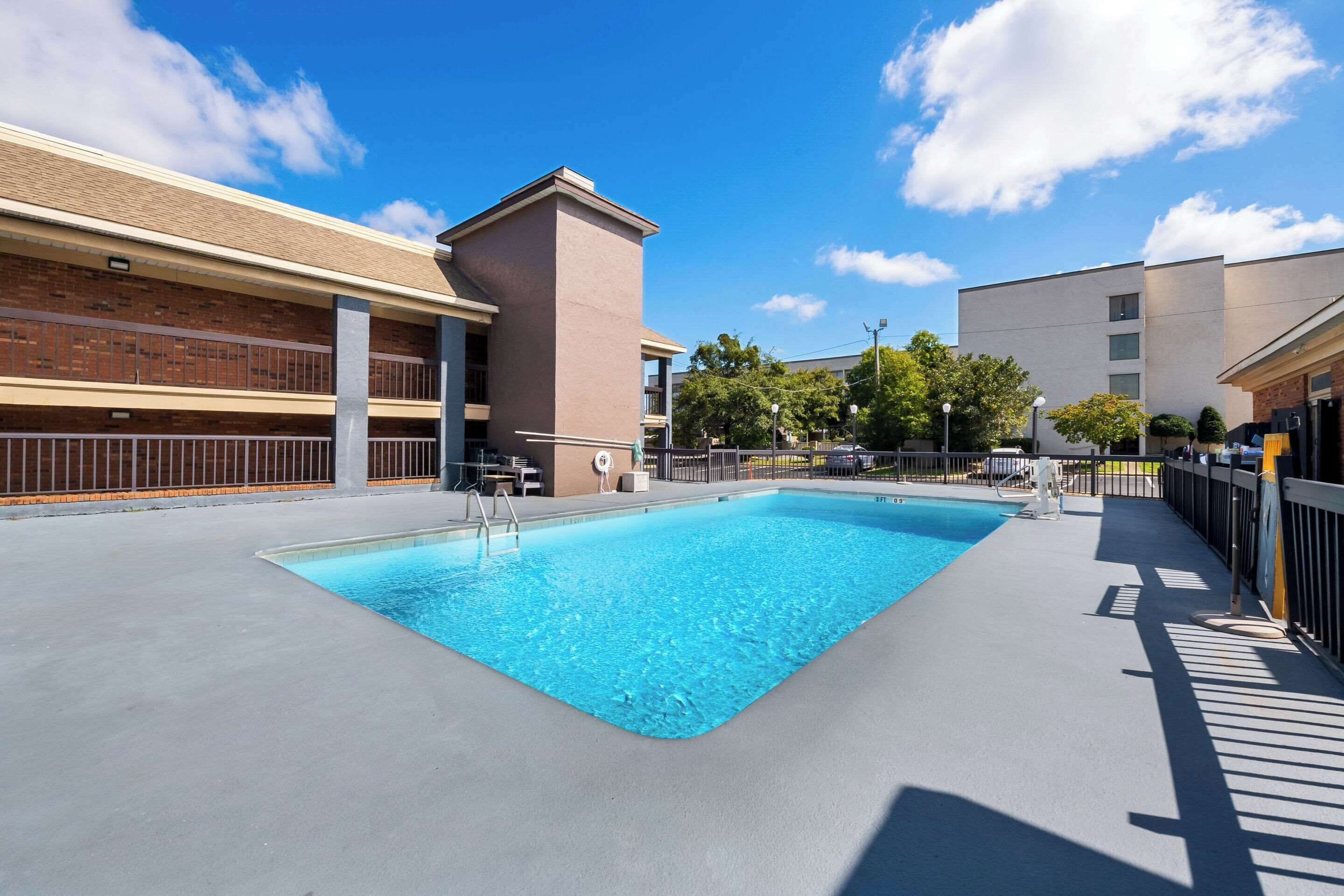 A rectangular swimming pool with a concrete deck, surrounded by a metal fence and a building with a brick facade.