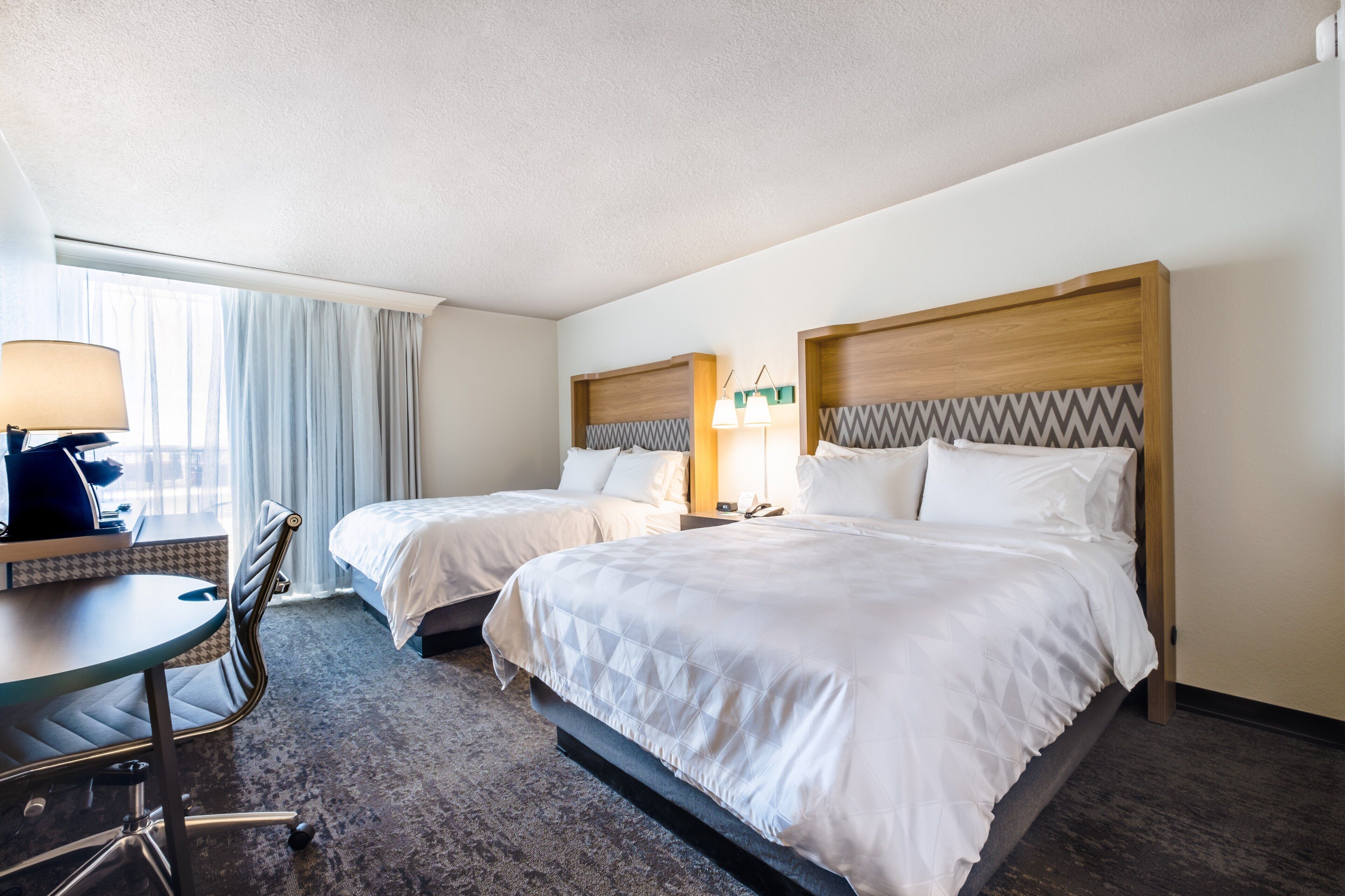 Une chambre d’hôtel avec deux lits, un bureau, une chaise et une fenêtre avec des rideaux.