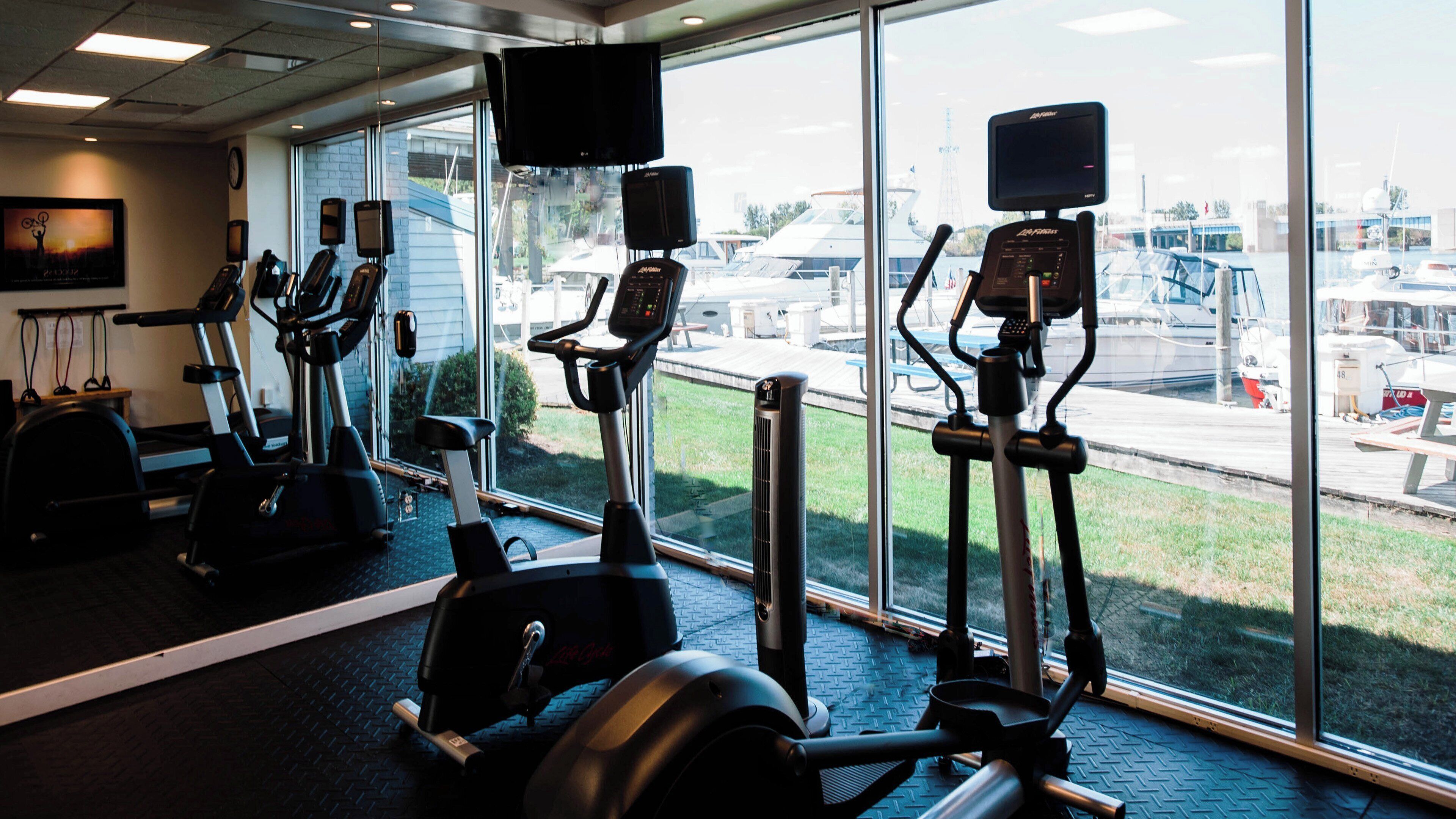 A gym with stationary bikes and a treadmill, large windows with a view of boats and a dock.