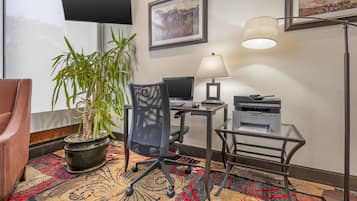 A hotel room with a desk, chair, computer, printer, potted plant, and a television mounted on the wall.