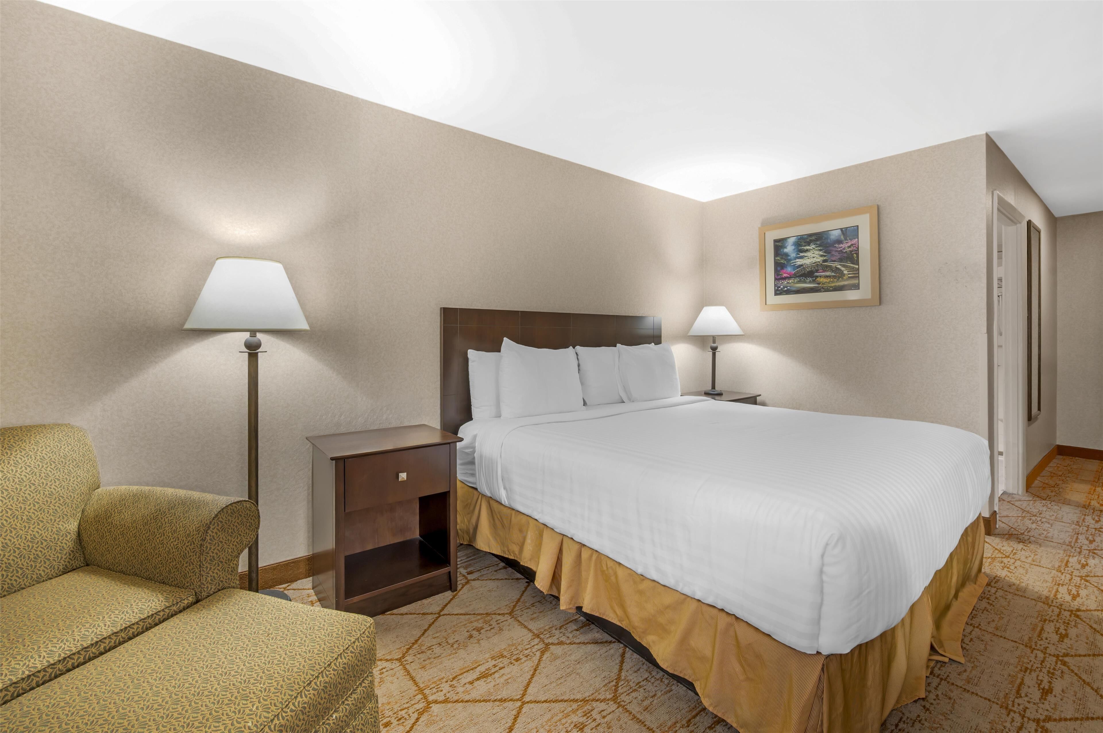 Habitación de hotel con cama, dos lámparas de noche, una mesita de noche, una silla y un cuadro en la pared.
