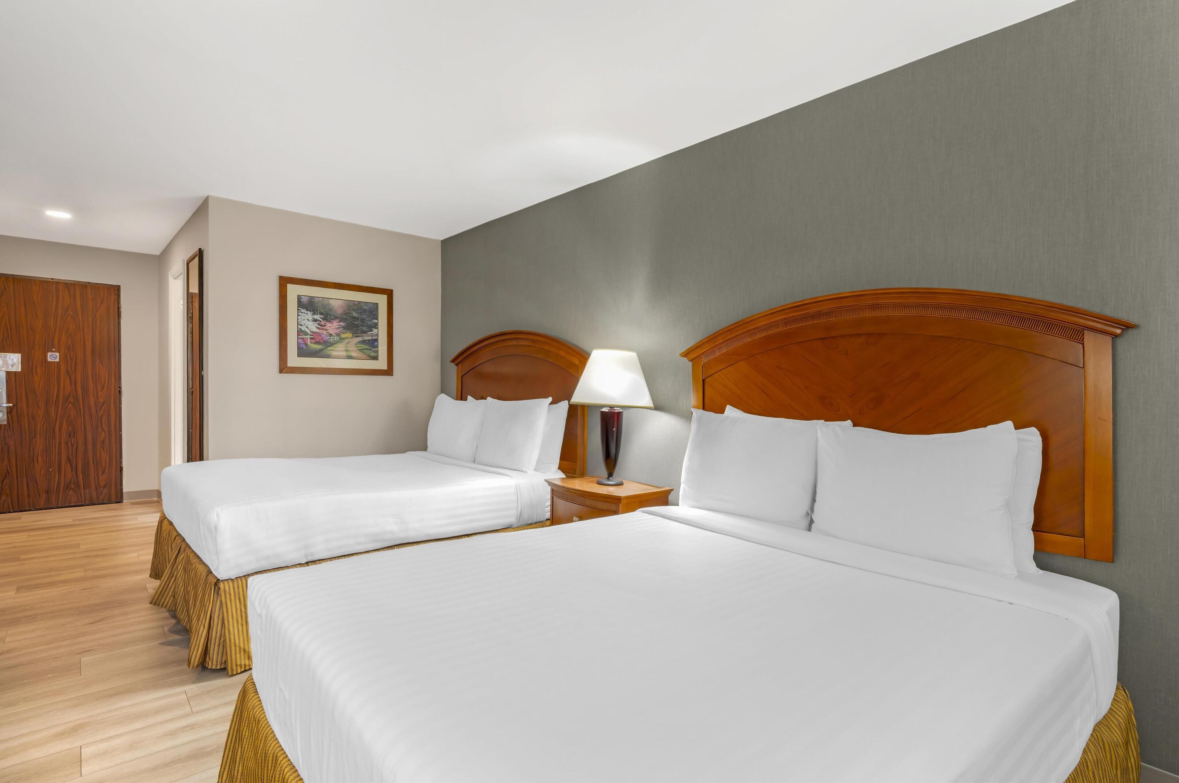 A hotel room with two single beds, wooden headboards, a nightstand with a lamp, and a framed picture on the wall.