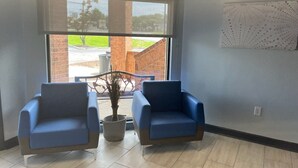Une pièce avec deux fauteuils bleus, une plante en pot et une fenêtre donnant sur un bâtiment en briques et sur la verdure environnante.