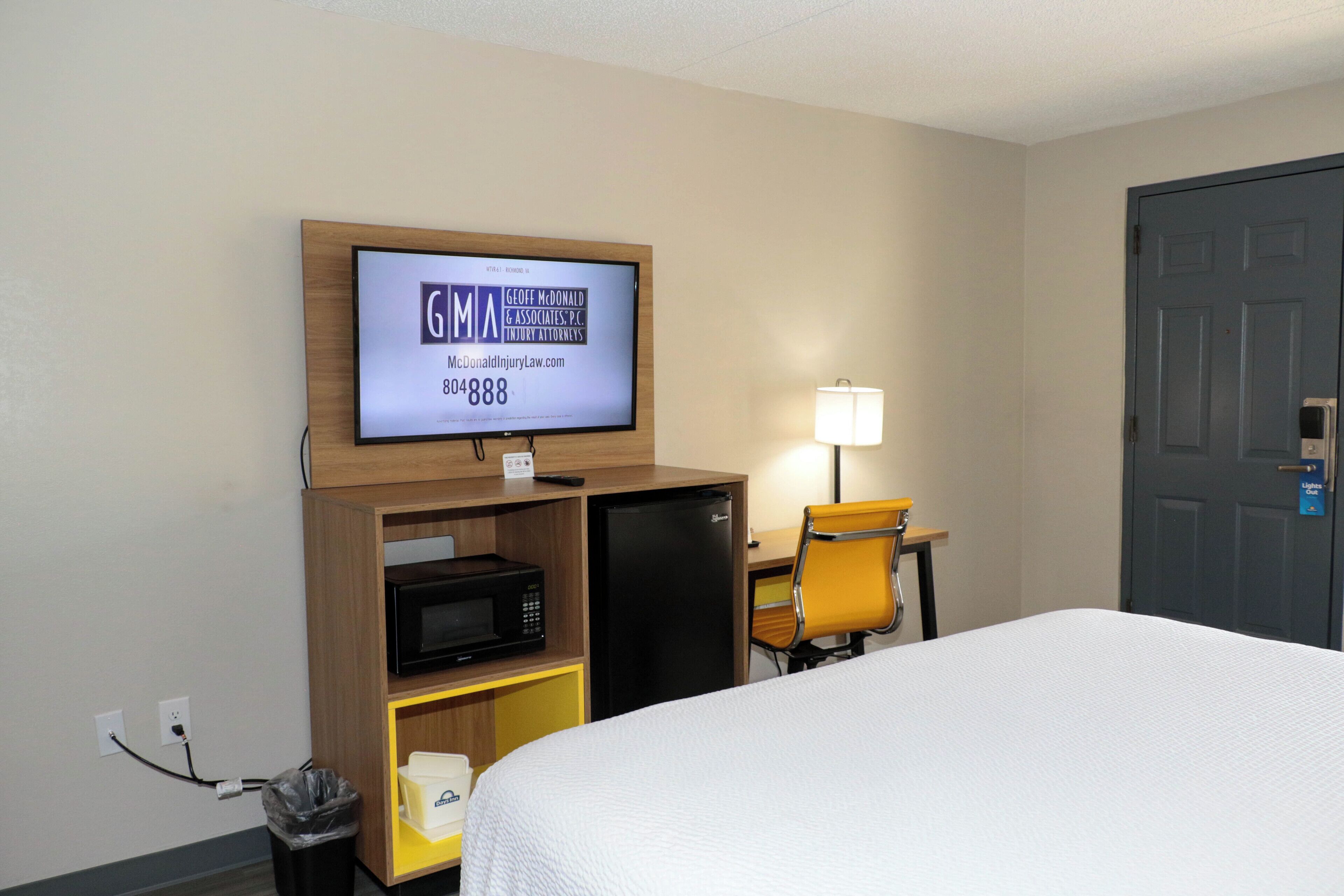 A hotel room with a bed, a television mounted on a wooden shelf, a desk with a chair, a lamp, and a door.