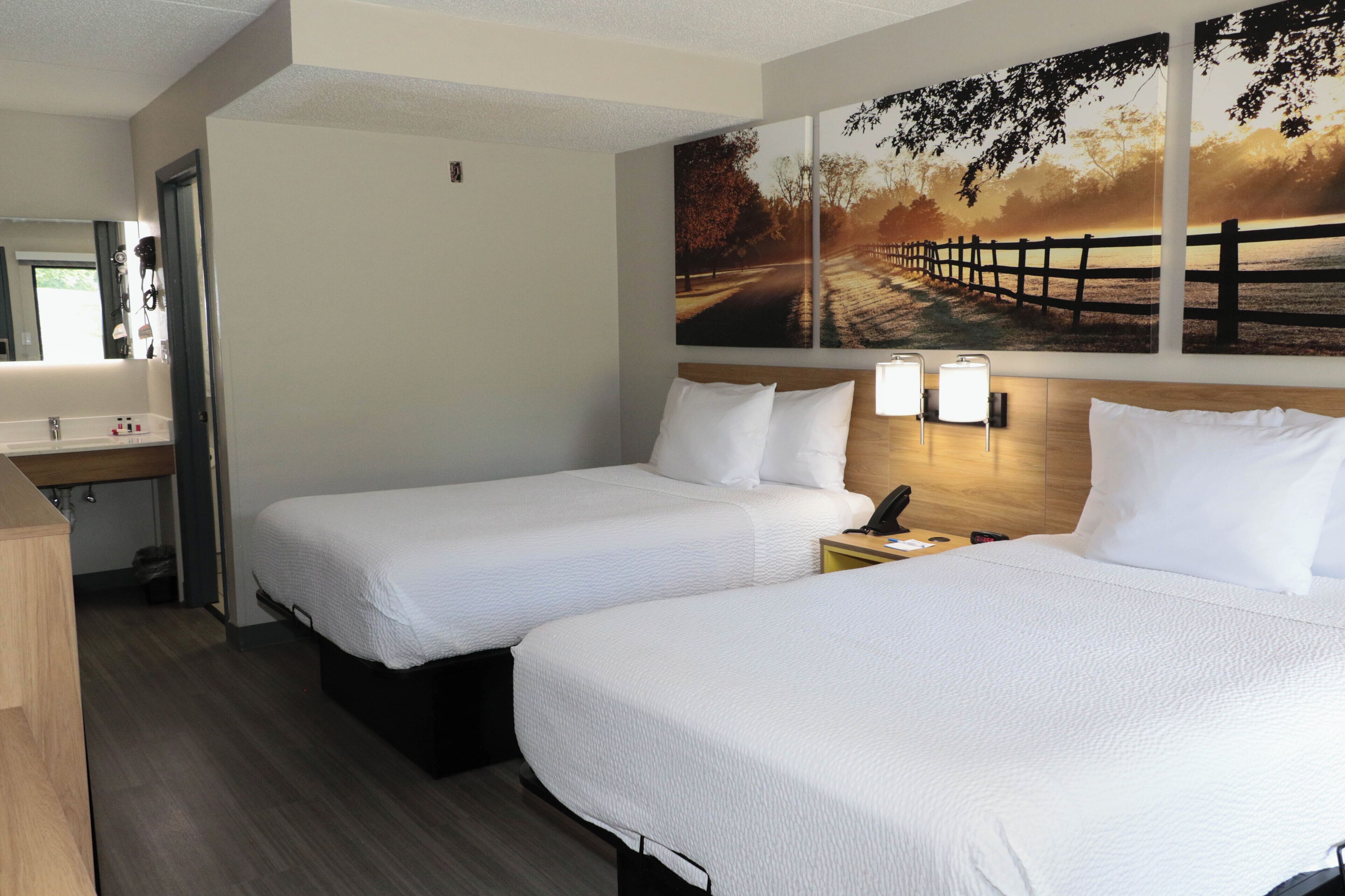 Une chambre d’hôtel avec deux lits, une tête de lit en bois, une table de chevet avec une lampe et un grand tableau représentant un paysage accroché au mur.