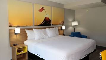 A hotel room with a large bed, two bedside tables, a nightstand, a desk, and a chair. There are two wall-mounted lamps and a painting of a sunset with a flag on the wall.
