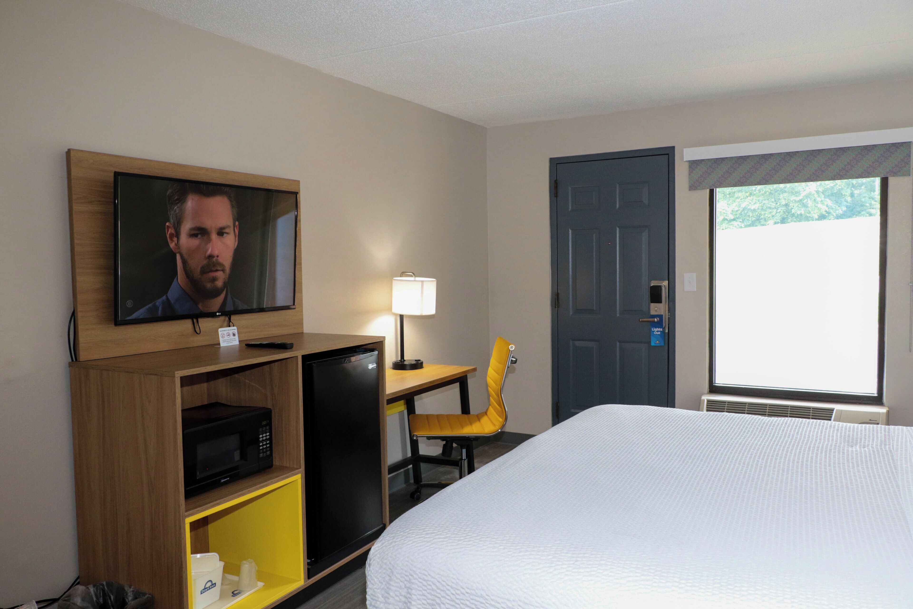 Habitación de hotel con una cama, un televisor montado en un estante de madera, un escritorio con una silla, una lámpara, un microondas y una puerta con ventanal.