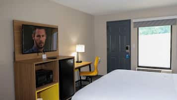 Ein Hotelzimmer mit einem Bett, einem an einer Holzwand montierten Fernseher, einem Schreibtisch mit Stuhl, einer Lampe, einer Mikrowelle und einer Tür mit Fenster.