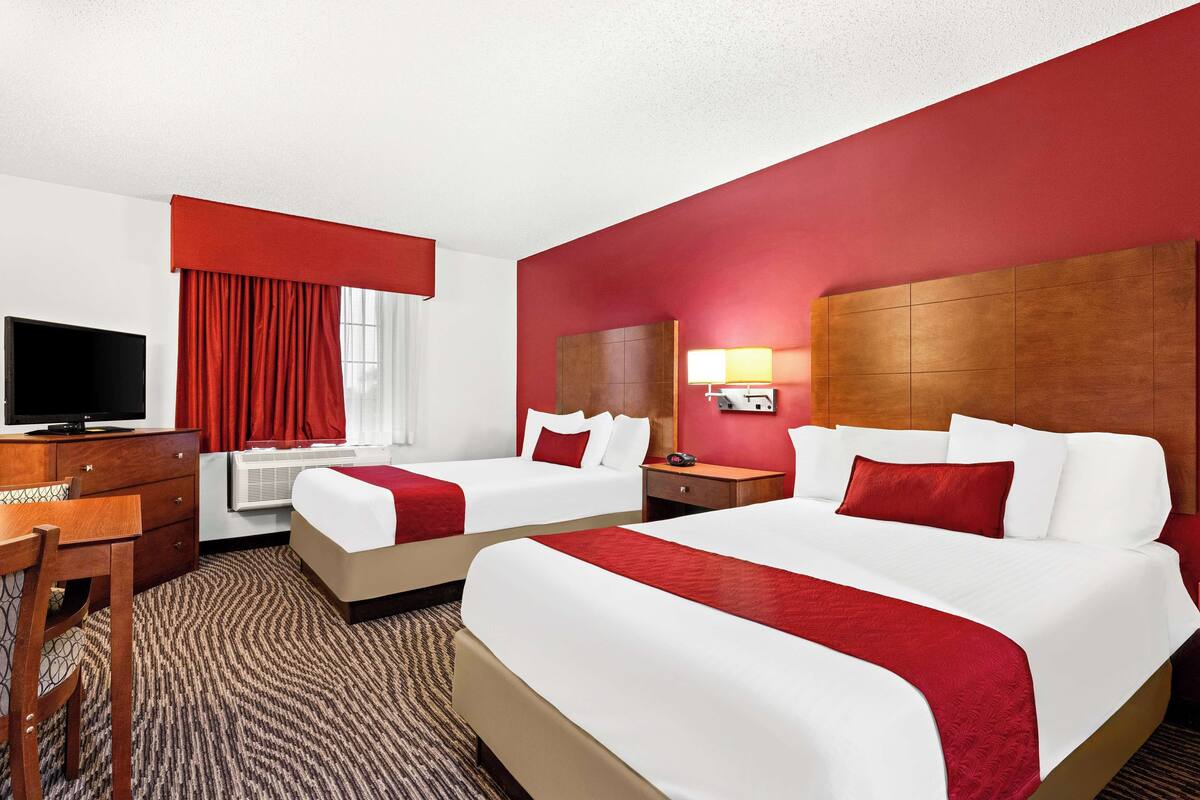 A hotel room with two beds, a desk, a television, and a window with red curtains.