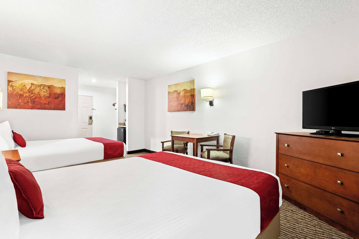 A hotel room with two beds, a television, a desk with chairs, and artwork on the walls.