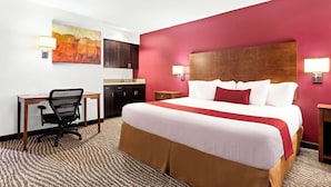 Una habitación de hotel con una cama grande, un escritorio con silla, una kitchenette pequeña y un cuadro en la pared.