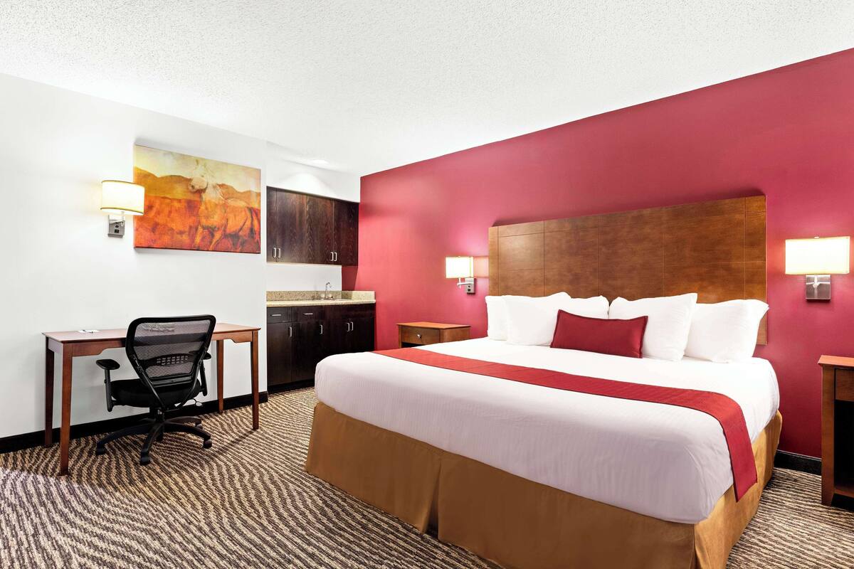 A hotel room with a large bed, a desk with a chair, a small kitchenette, and a painting on the wall.