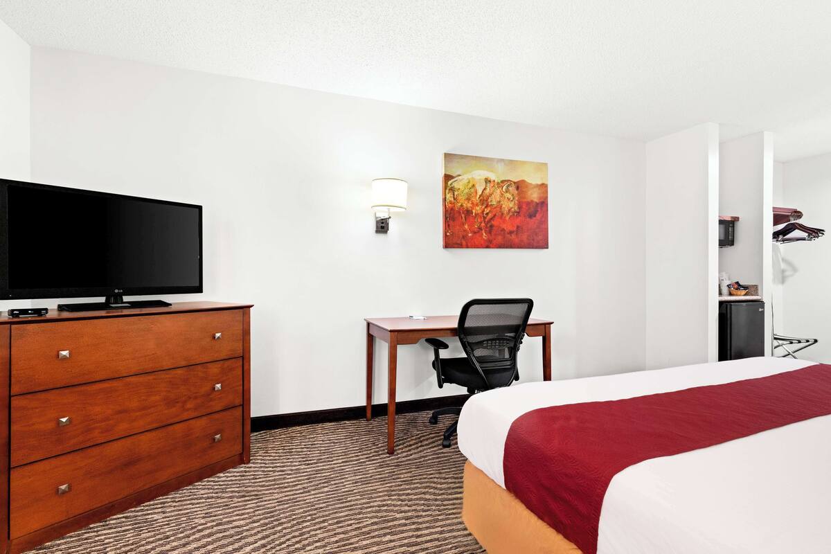 A hotel room with a bed, a desk with a chair, a television on a dresser, and a painting on the wall.