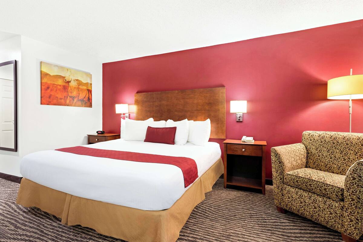 A hotel room with a large bed, a red accent wall, a patterned armchair, and a painting on the wall.